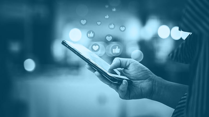 Hand hält ein Smartphone, aus dem Display steigen Social Media Symbole wie Seifenblasen