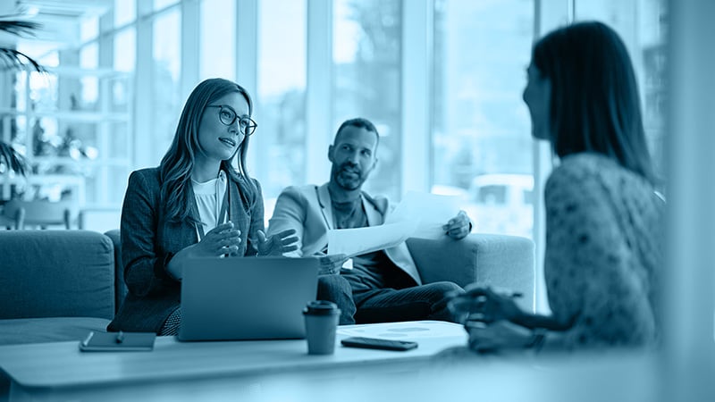 Drei Personen im Business-Meeting, zwei sitzen auf dem Sofa mit Laptop und Dokumenten, eine Person gegenüber – Symbol für strategische Gespräche bei PR-Redaktionstouren